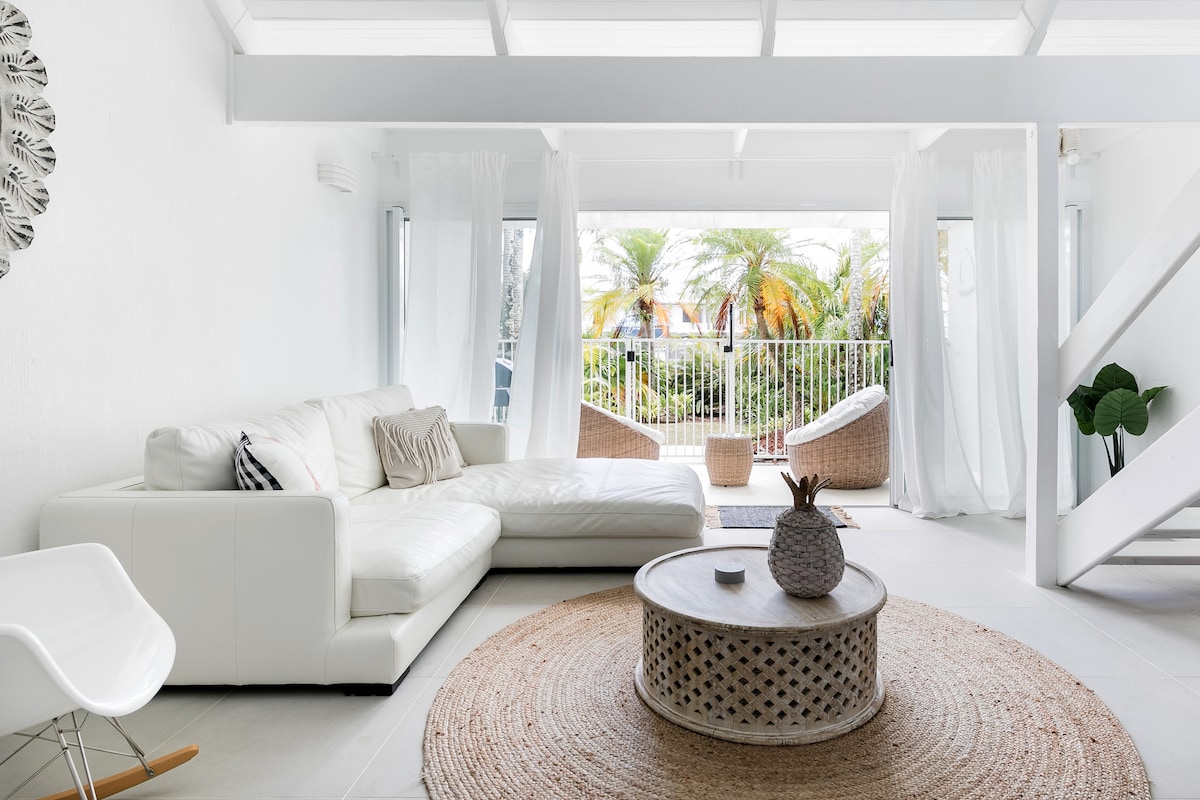 A light-filled living area features a white sectional sofa positioned near large windows with sheer drapes. A round woven rug lies beneath a decorative coffee table, while a plant and basket chairs are visible through the glass doors leading to a balcony with garden views.