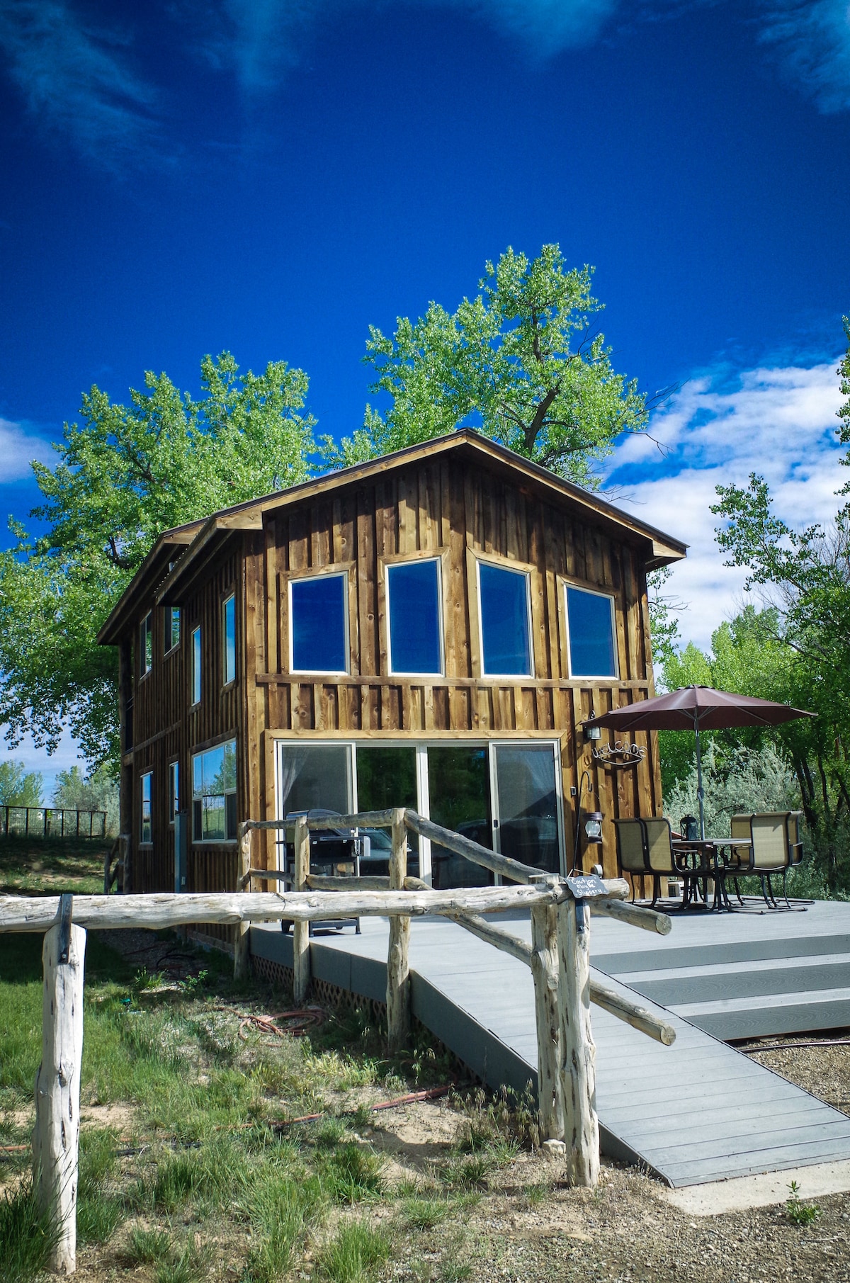 The newly built house features rustic wooden siding and large windows, allowing for ample natural light. A covered deck is visible with an outdoor table and chairs under a shade umbrella, providing a serene spot to enjoy the surrounding landscape.