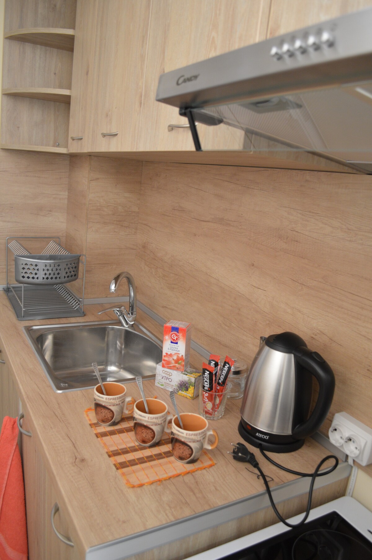 A well-equipped kitchen features light wood cabinetry and a stainless steel kettle. A sink and cooking surface are visible, alongside neatly arranged mugs on an orange dish towel. Essential items, including coffee packets and a carton, are positioned near the kettle.