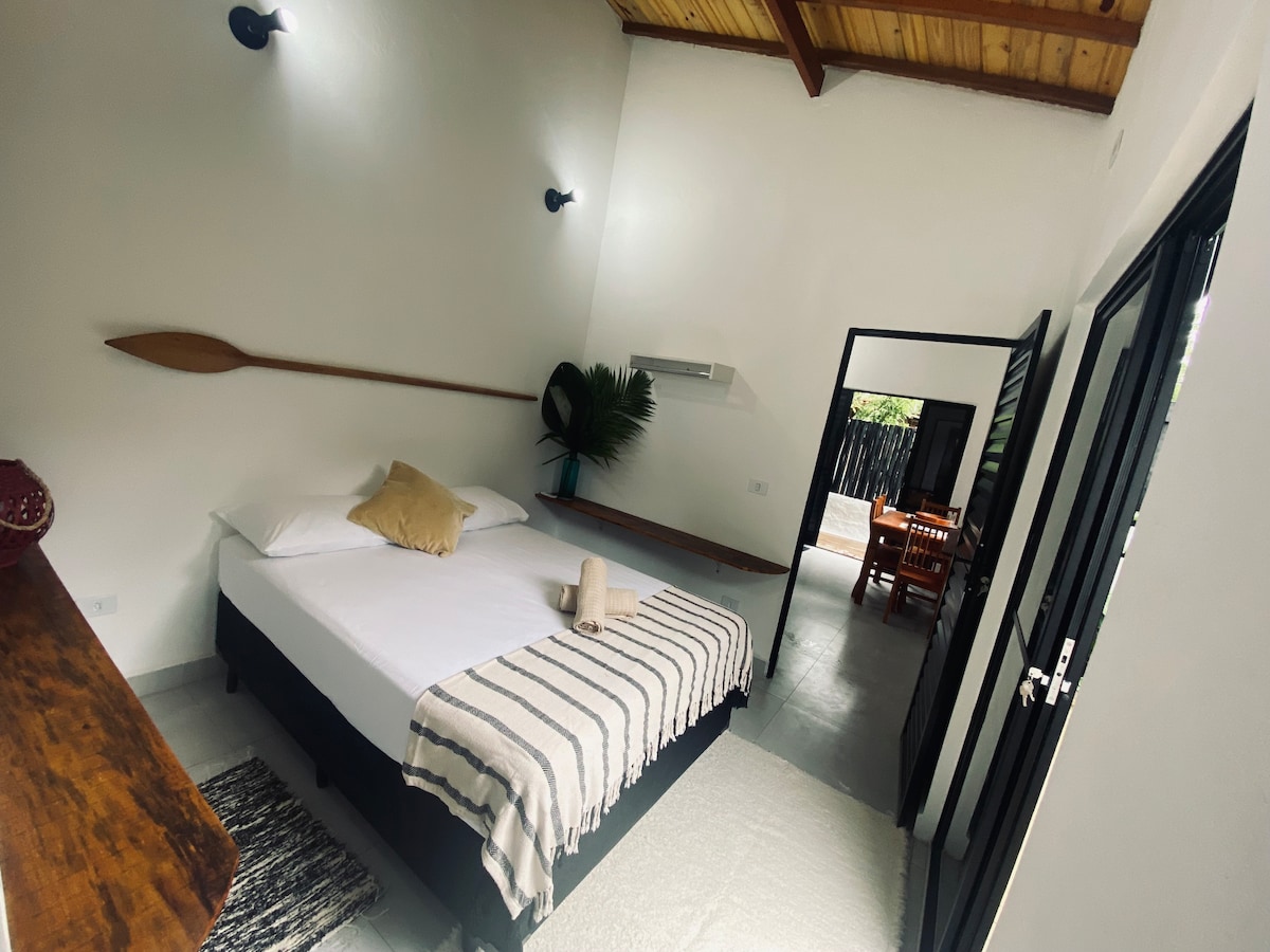 A bright sleeping area features a spacious bed with a light-colored blanket and a decorative pillow. Wooden beams line the ceiling, and a large paddle adorns the wall. A breeze is invited through an open doorway leading to a dining area visible in the background.
