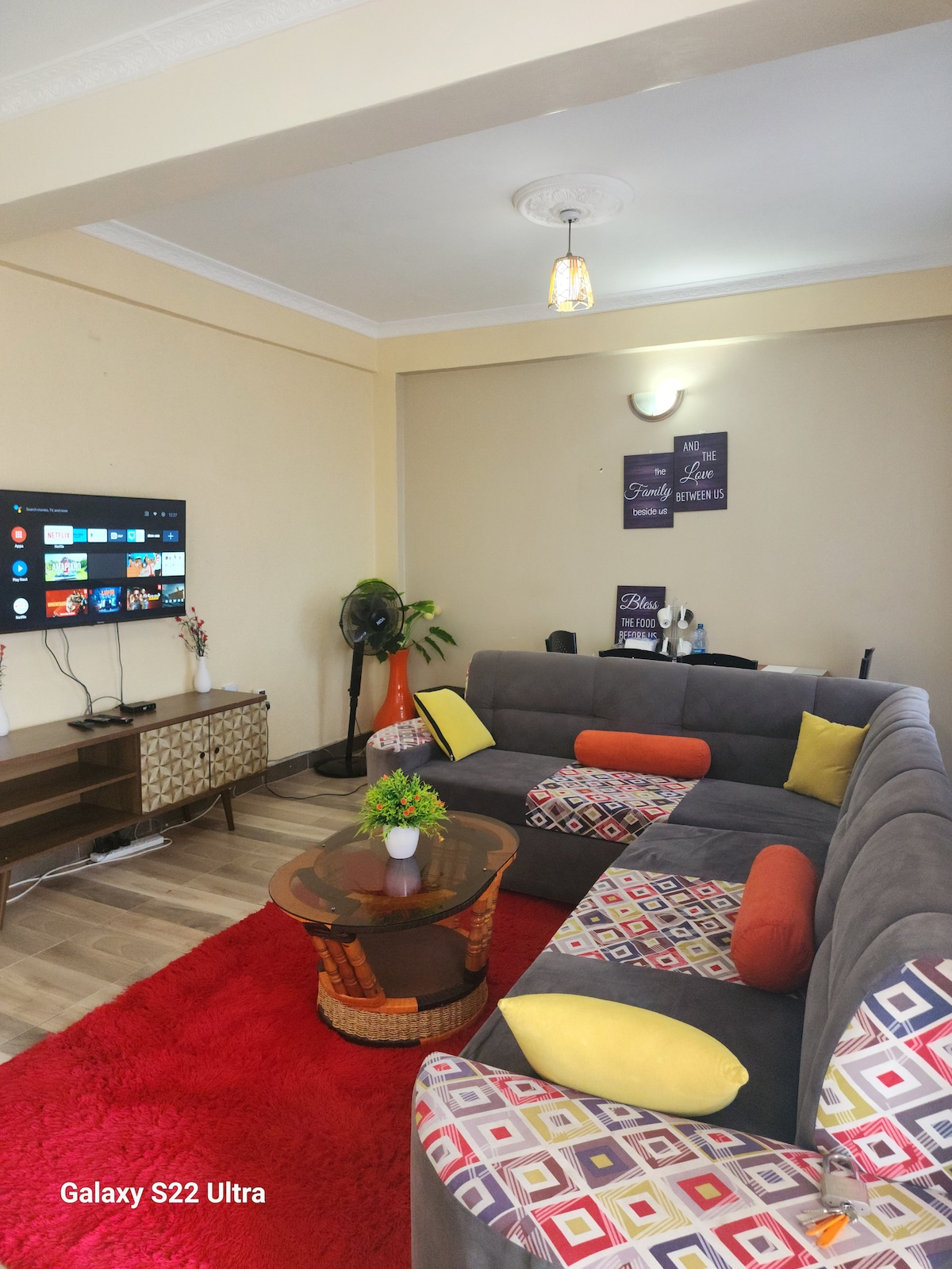 A spacious living area features a large gray sectional sofa adorned with colorful patterned cushions and orange accent pillows. A glass coffee table rests on a vibrant red rug, while a flat-screen TV is mounted on the wall. A small fan and decorative wall art are also visible.