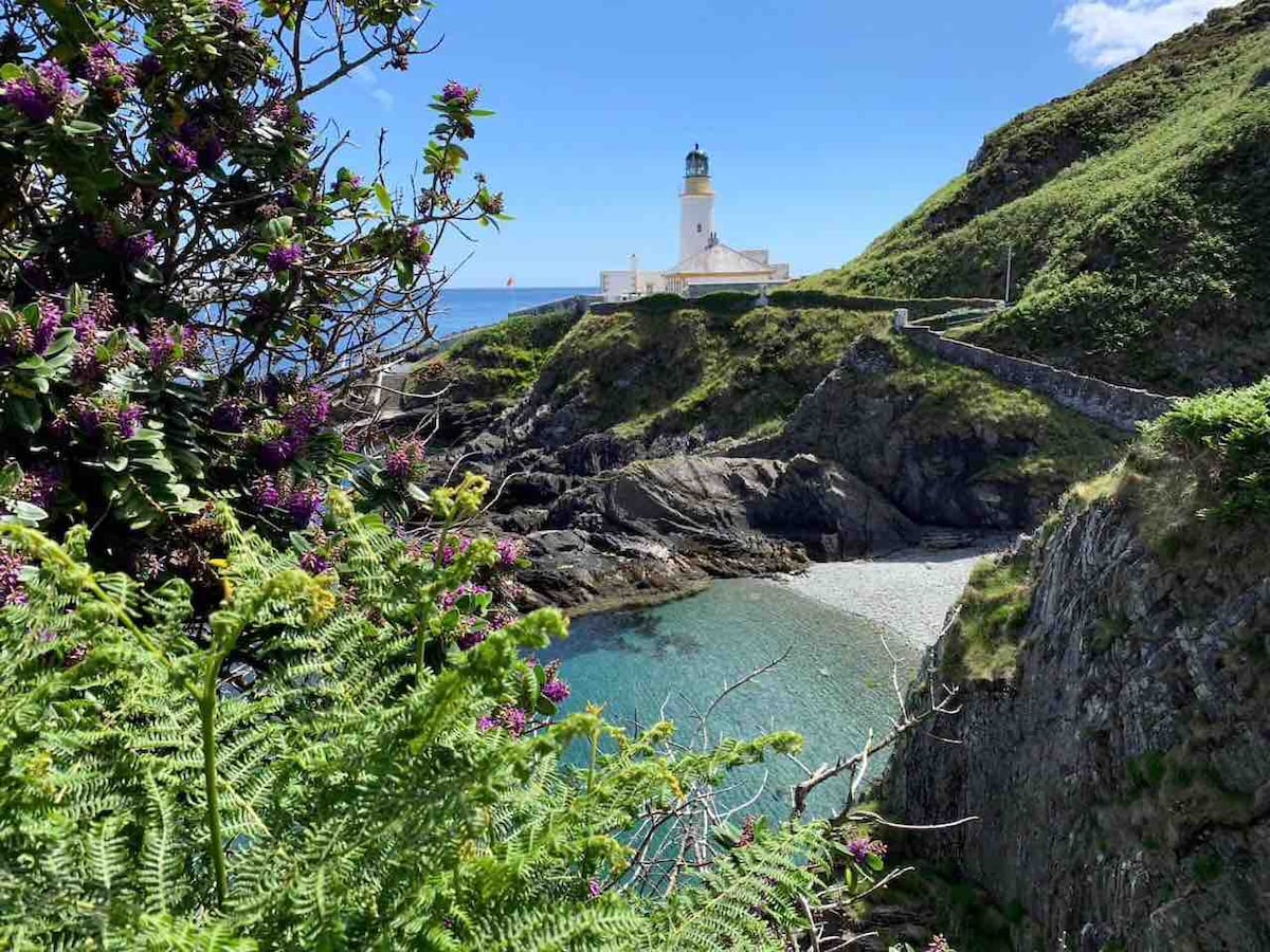 The image showcases a historic lighthouse nestled on a coastal hill, surrounded by lush greenery and colorful flowers. A tranquil cove is visible in the foreground, with clear blue waters and rocky shoreline, while the sky above is bright and clear.