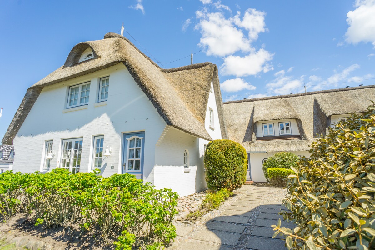 A charming building with a thatched roof features white walls and multiple windows. Lush greenery and a stone pathway lead to the entrance, creating a welcoming exterior against a clear blue sky.