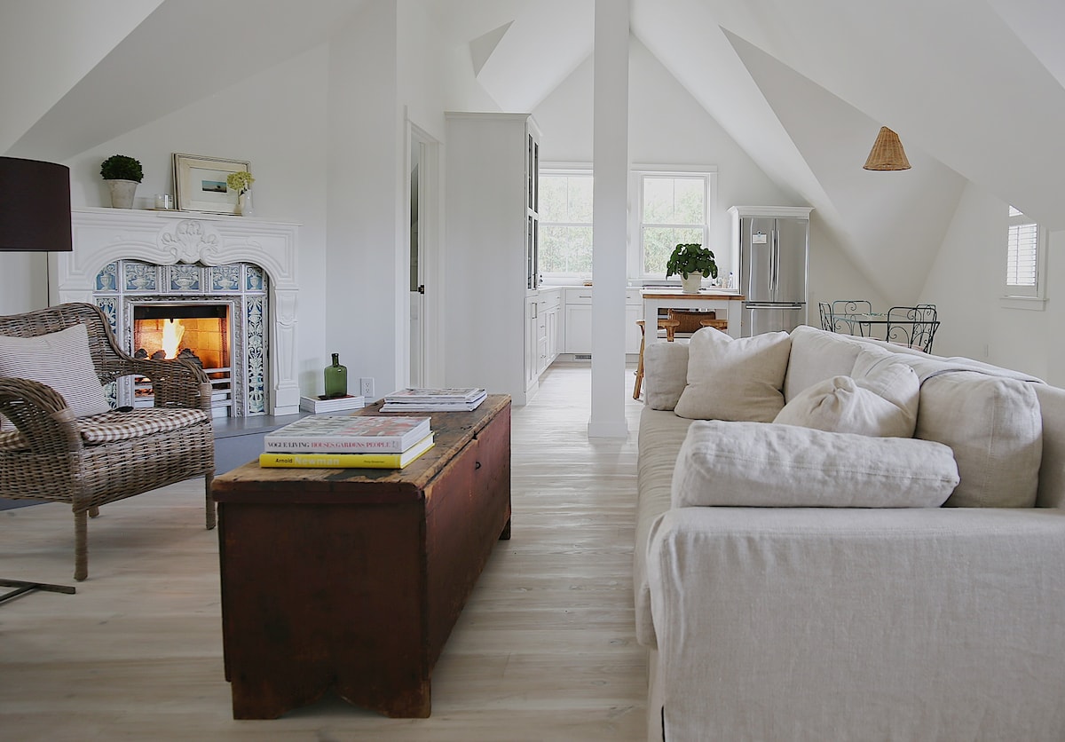 A bright, open living space features vaulted ceilings and light-colored walls. A cozy seating area includes a plush sofa and a wicker chair near a decorative fireplace. Natural light floods the room, illuminating a vintage coffee table and an elegant dining area in the background.