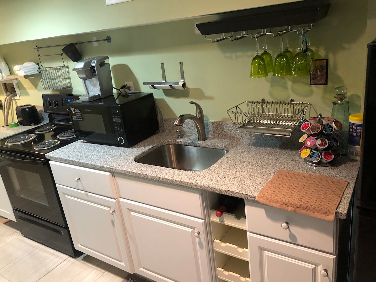 The kitchen area features a well-maintained countertop with a stainless steel sink and a microwave. Below, ample cabinetry provides storage. Hanging above the counter, decorative green glasses are arranged. A coffee pod holder is visible, and appliances include a stove and an oven.