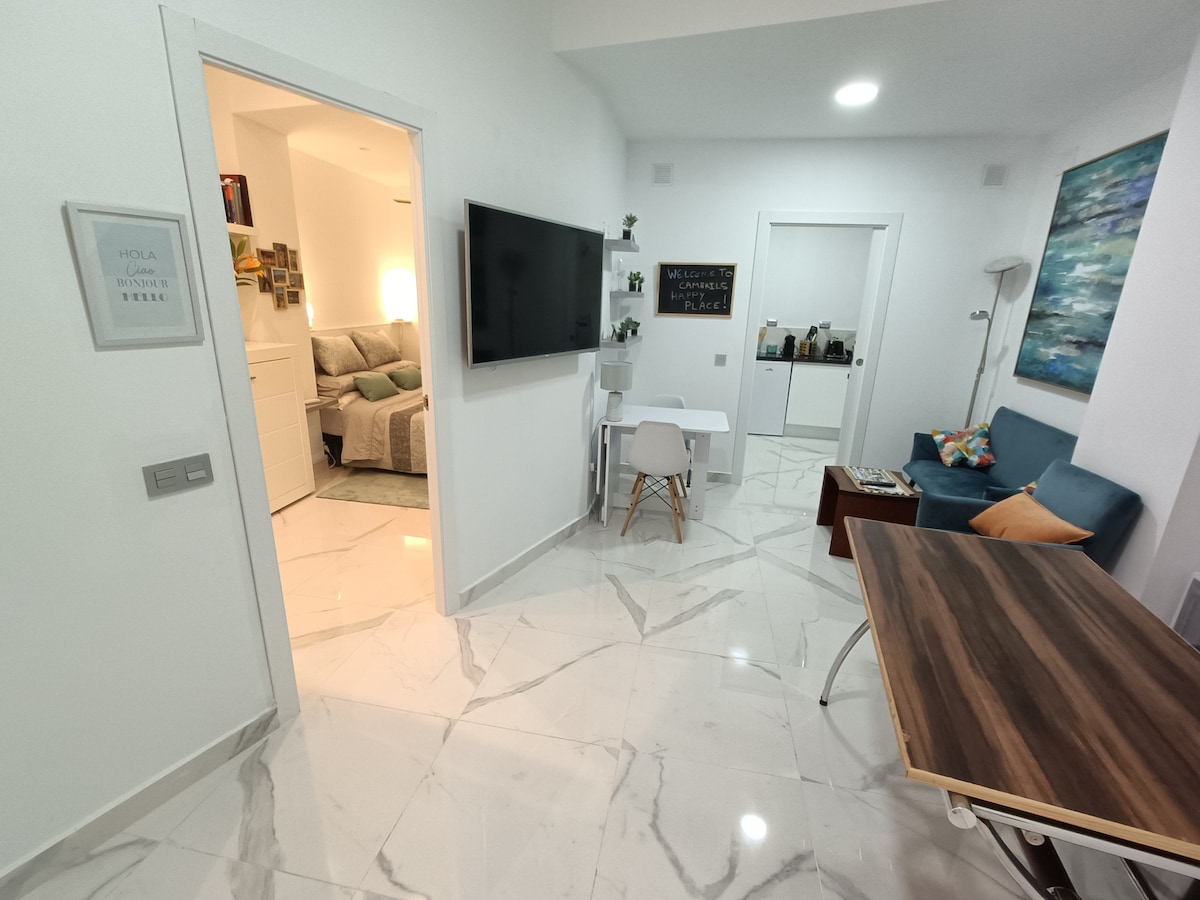 A well-lit entrance area is visible, featuring glossy porcelain tiles. A modern television is mounted on the wall, while a sleek white desk and chair provide a workspace. A cozy seating area with a blue sofa and a wooden dining table creates a welcoming environment.