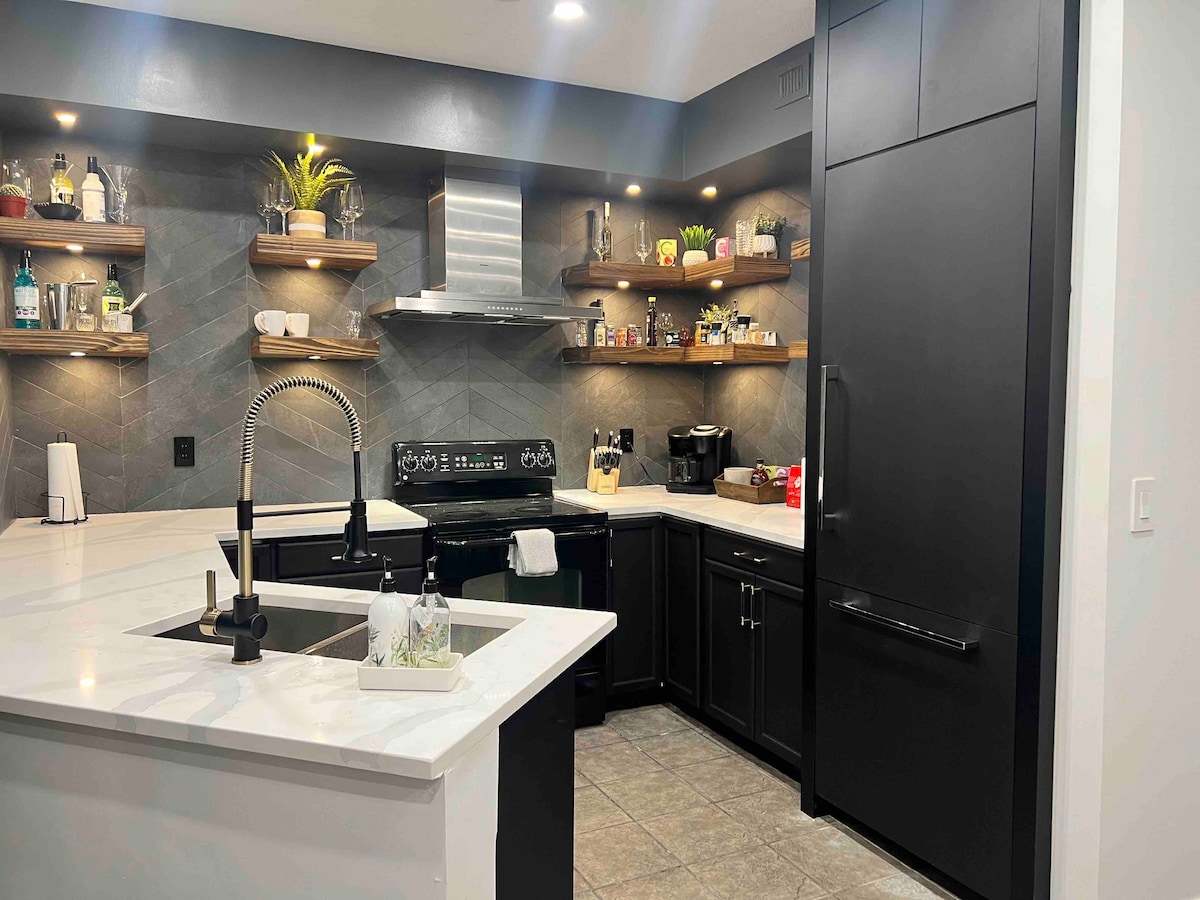 A modern kitchen featuring sleek black cabinets and white countertops is illuminated by under-cabinet lighting. Open shelving displays various decorative items and kitchen essentials. A stainless steel refrigerator stands prominently, and a dual-basin sink with a stylish faucet completes the space.