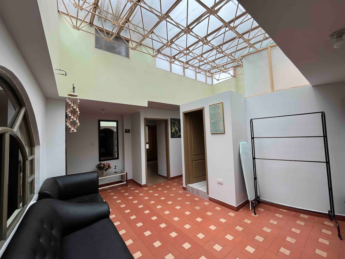 The interior space is well-lit by a skylight, revealing a spacious area with a black sofa and terracotta tiled flooring. Walls are painted in light colors, and doorways lead to other rooms. A small decorative plant adds a touch of greenery to the setting.