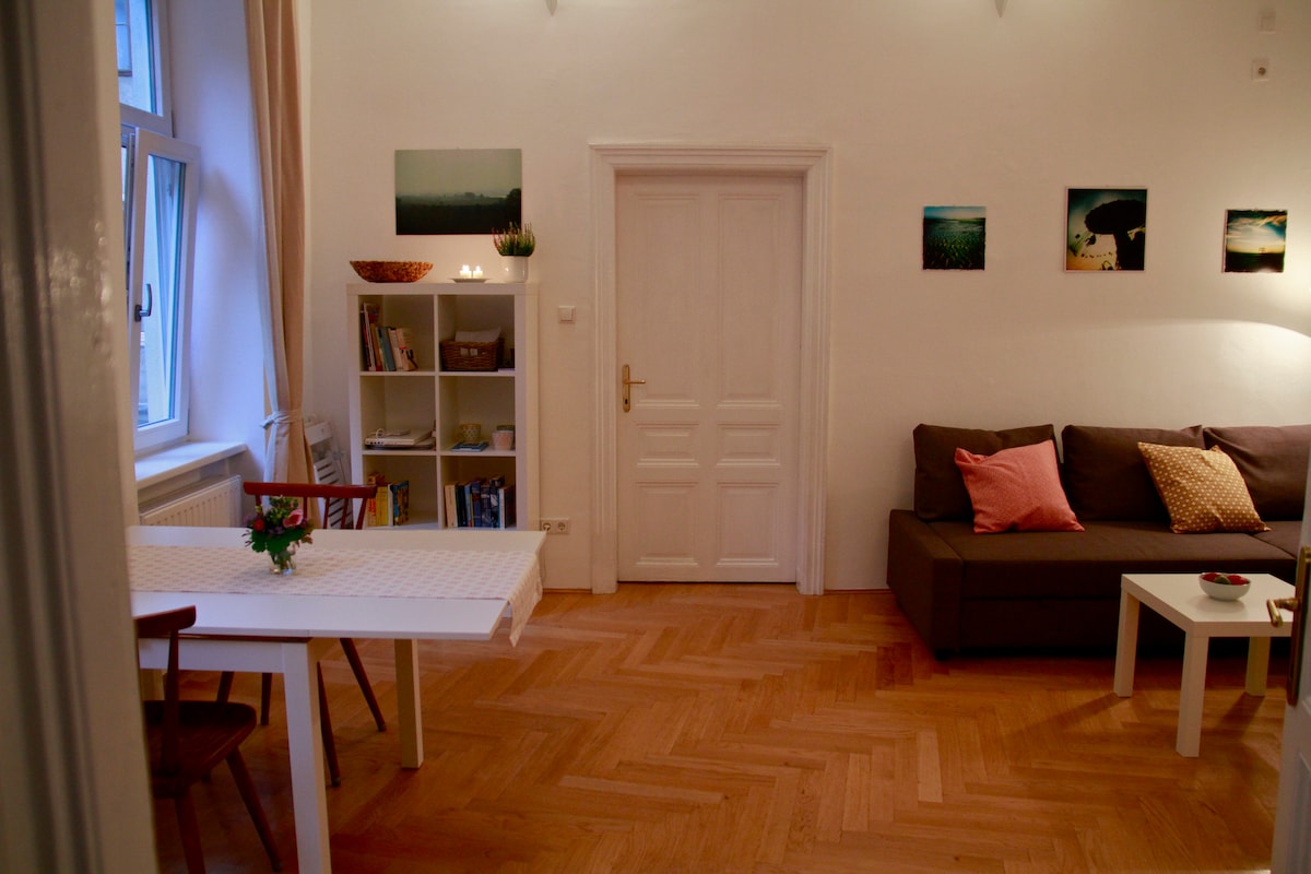 A spacious living area features a comfortable sofa adorned with decorative pillows. A dining table, set for six, is positioned nearby. A bookshelf holds various items, and a small table is placed in front of the sofa. Soft natural light enters through a window, enhancing the cozy atmosphere.