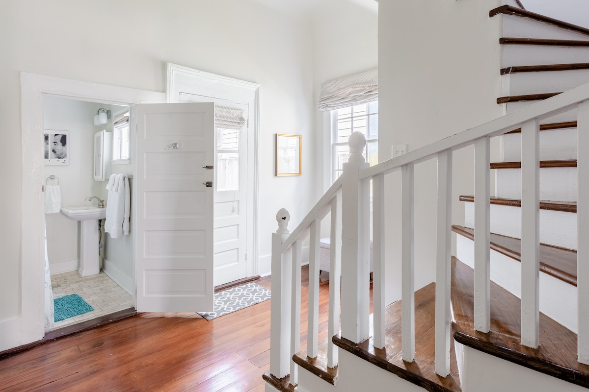 An airy entryway features a staircase with polished wooden treads leading to the upper level. A doorway on the left opens to a bathroom, while natural light fills the space through the windows, illuminating light-colored walls and rustic plank flooring.