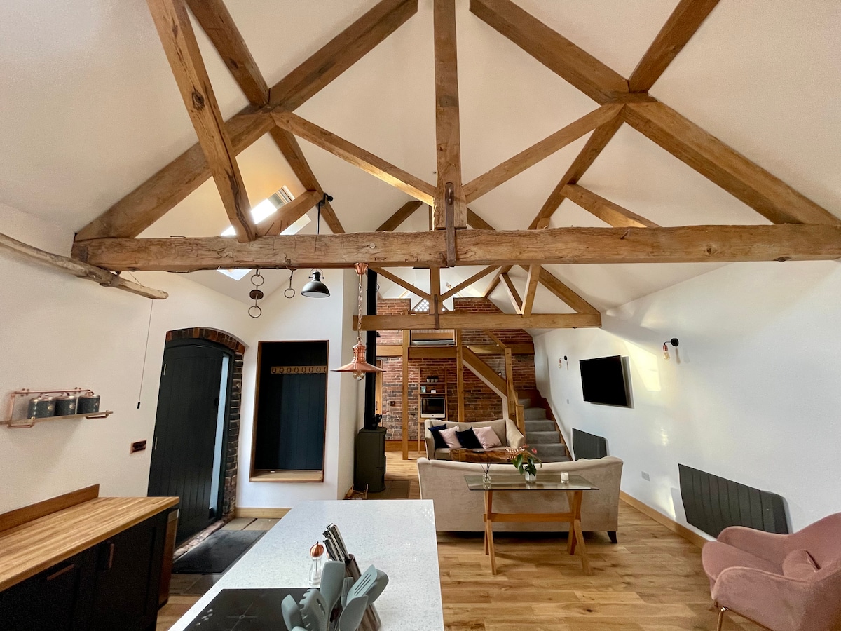 The interior features a high ceiling with exposed wooden beams, creating an open and airy atmosphere. A central kitchen island with a sleek countertop is complemented by modern lighting fixtures. A comfortable seating area and exposed brick walls add warmth to the space.