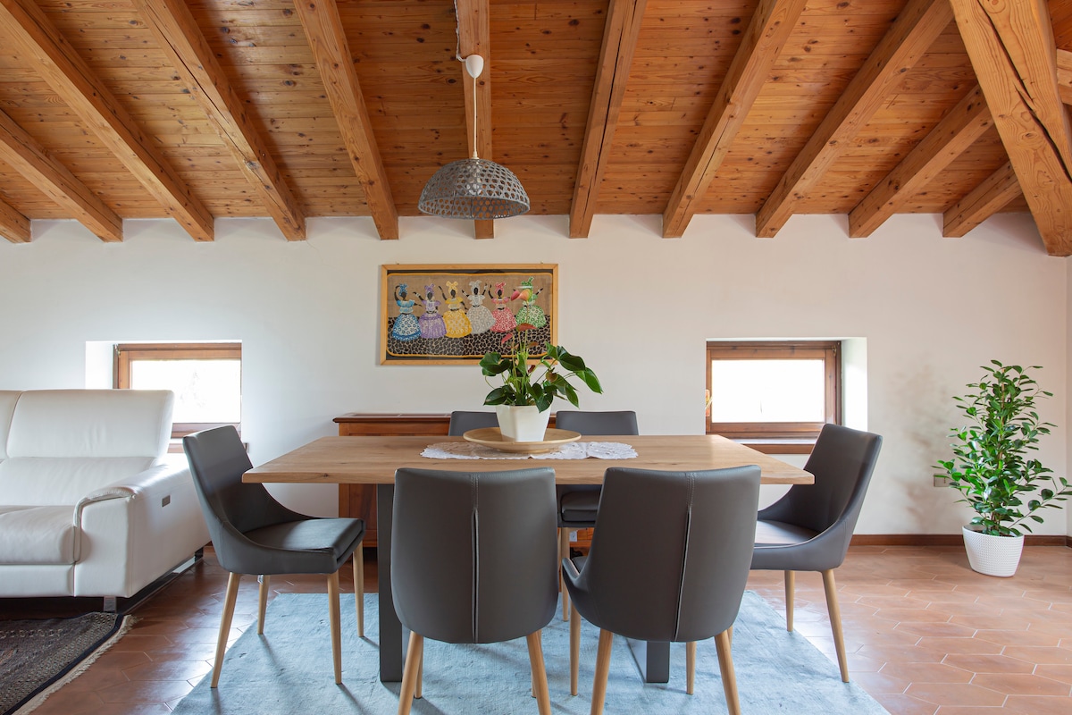 An inviting dining area is featured, furnished with a wooden table surrounded by six grey chairs. Exposed wooden beams create a warm ceiling. Natural light enters through two windows, complemented by a decorative artwork on the wall and a potted plant in the corner.