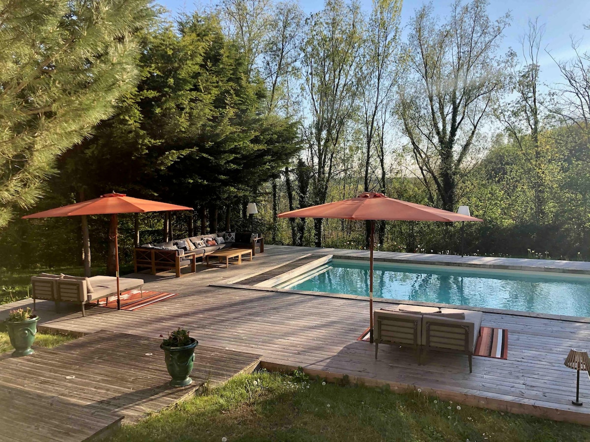 A well-maintained outdoor area features a spacious deck surrounding a heated swimming pool. Comfortable lounge chairs provide shade under large umbrellas, while lush greenery and trees create a serene backdrop. A seating arrangement is visible for relaxation beside the pool.