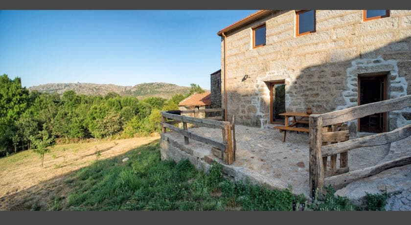 An exterior view of a stone house is presented, showcasing a wooden fence surrounding a patio area. A sturdy picnic table is positioned on the stone floor, and lush greenery extends toward the mountain backdrop, under a clear blue sky.