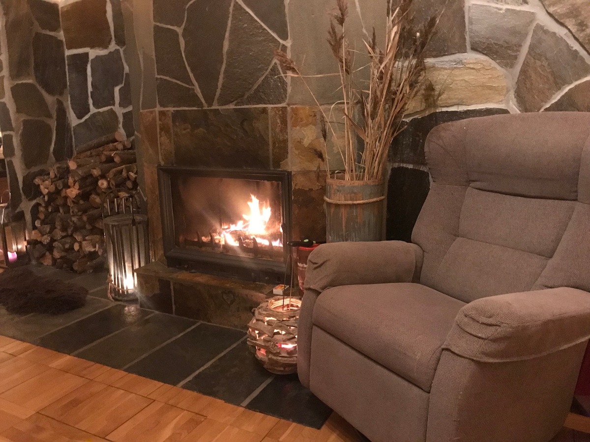 A cozy fireplace is framed by natural stone, providing warmth and ambiance. A comfortable armchair sits close by, accompanied by a decorative lantern and a stack of firewood. Subtle greenery adds a touch of nature, creating a welcoming space for relaxation.