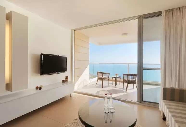 A modern living space features a flat-screen television mounted on a white unit. A glass door opens to a balcony, where two woven chairs and a small table overlook a scenic view. Natural light floods the room through sheer curtains.