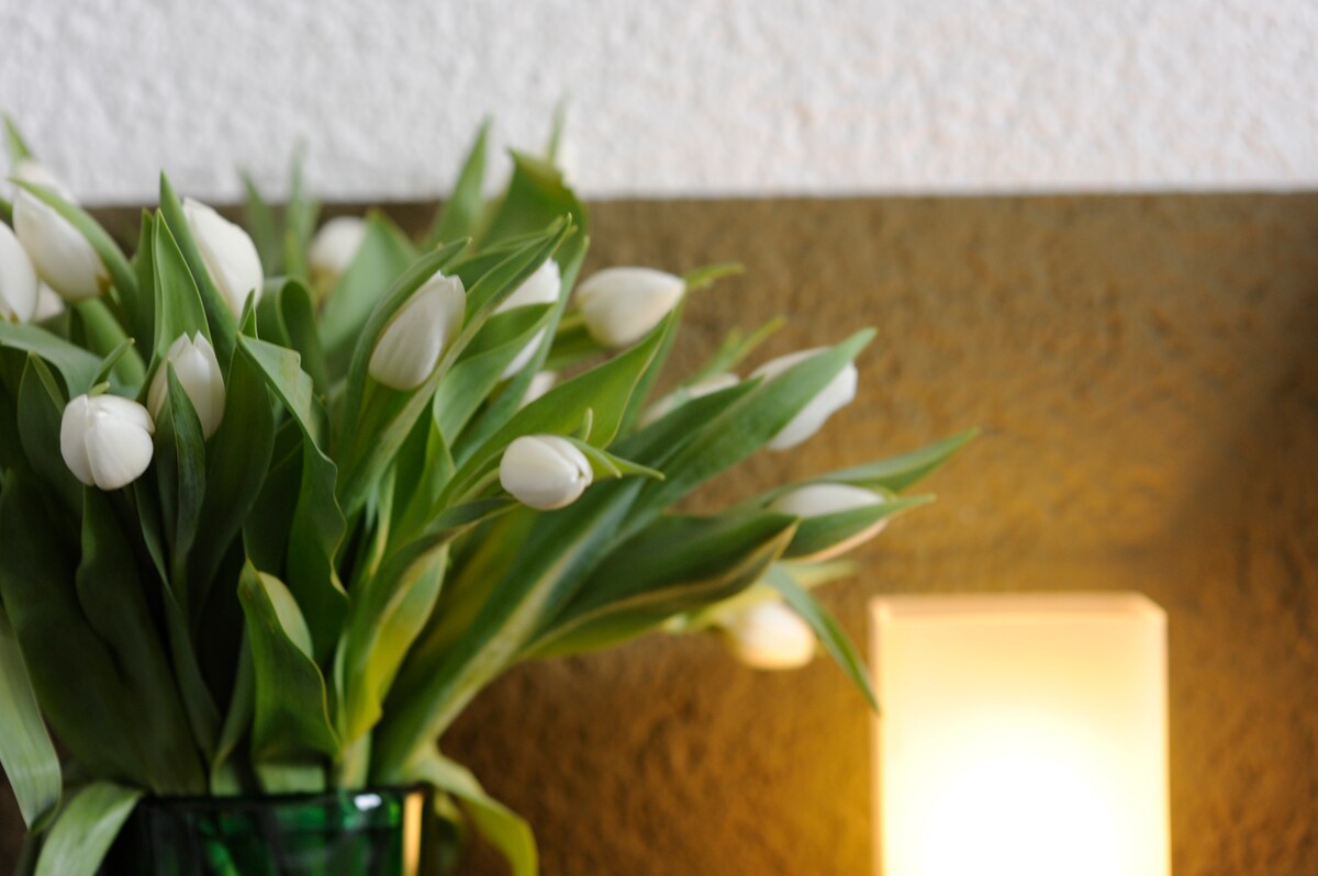A bouquet of white tulips is arranged in a green glass vase, positioned against a textured wall. A soft, warm light emanates from a nearby lamp, creating a serene and inviting ambiance.