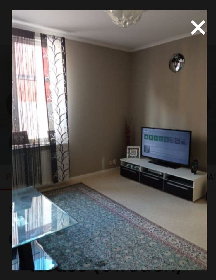A well-appointed living area is showcased, featuring a modern television on a sleek black entertainment unit. Natural light filters through sheer curtains, illuminating a decorative area rug that adds warmth to the neutral-toned room.