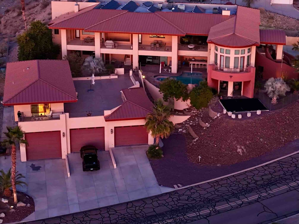 An aerial view of a modern house features a multi-level design with a prominent circular turret. The property includes a spacious driveway with three garage spaces, a large patio area, and a pool situated in a lush landscaped environment.