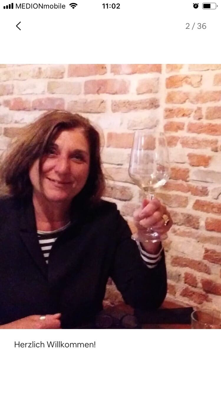 A person is seated at a table, holding a wine glass. The background features a rustic brick wall, adding warmth to the atmosphere. Subtle details like the striped shirt and the soft lighting enhance the inviting setting.