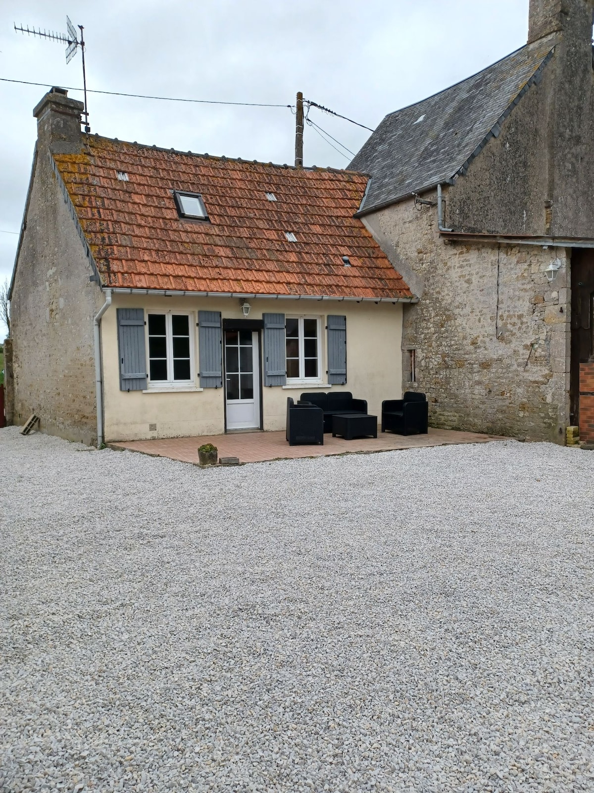 The exterior of a small independent house features a gravel courtyard and a comfortable seating area with dark chairs. The structure showcases a sloped roof with a mix of shingles and stone accents, framed by white shutters and windowpanes. A nearby building is visible, adding to the rural charm.