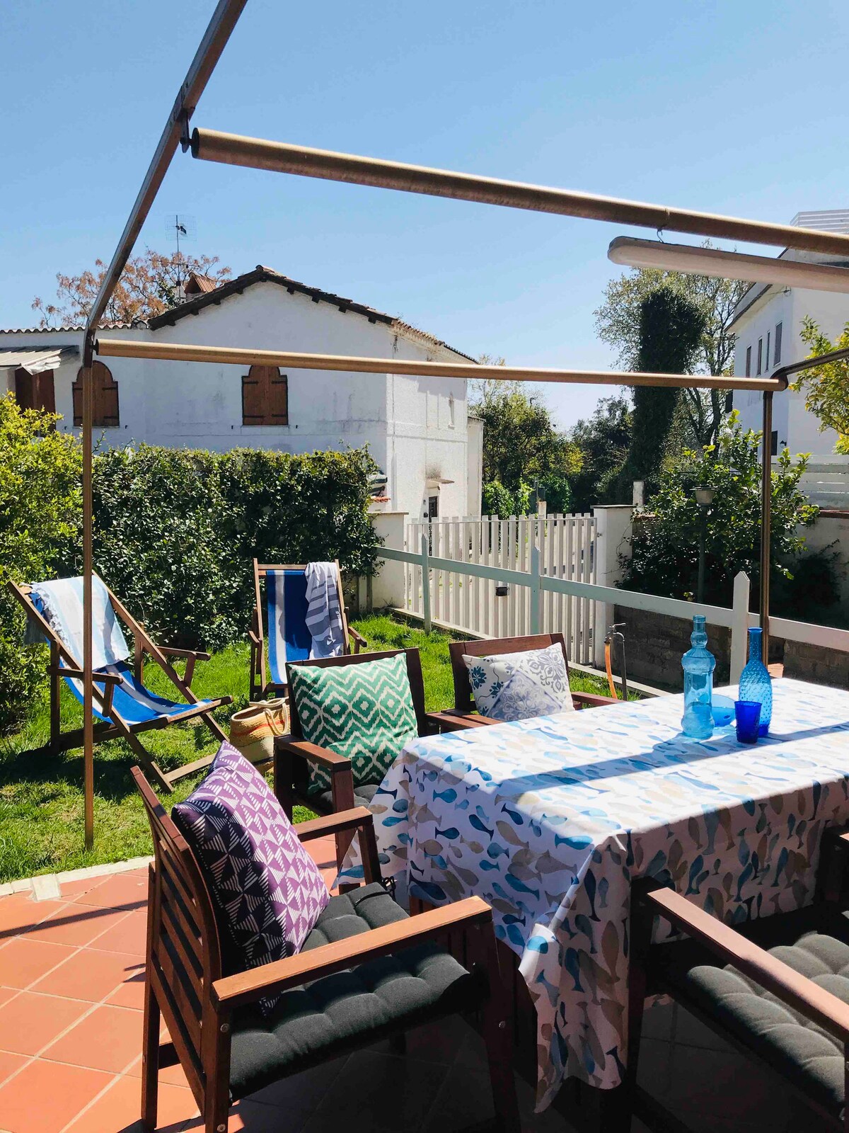 An outdoor dining area is presented, featuring a table adorned with a patterned tablecloth and surrounded by several wooden chairs. A sunny garden is visible in the background, complemented by green shrubs and a white picket fence. The sky is clear and blue.