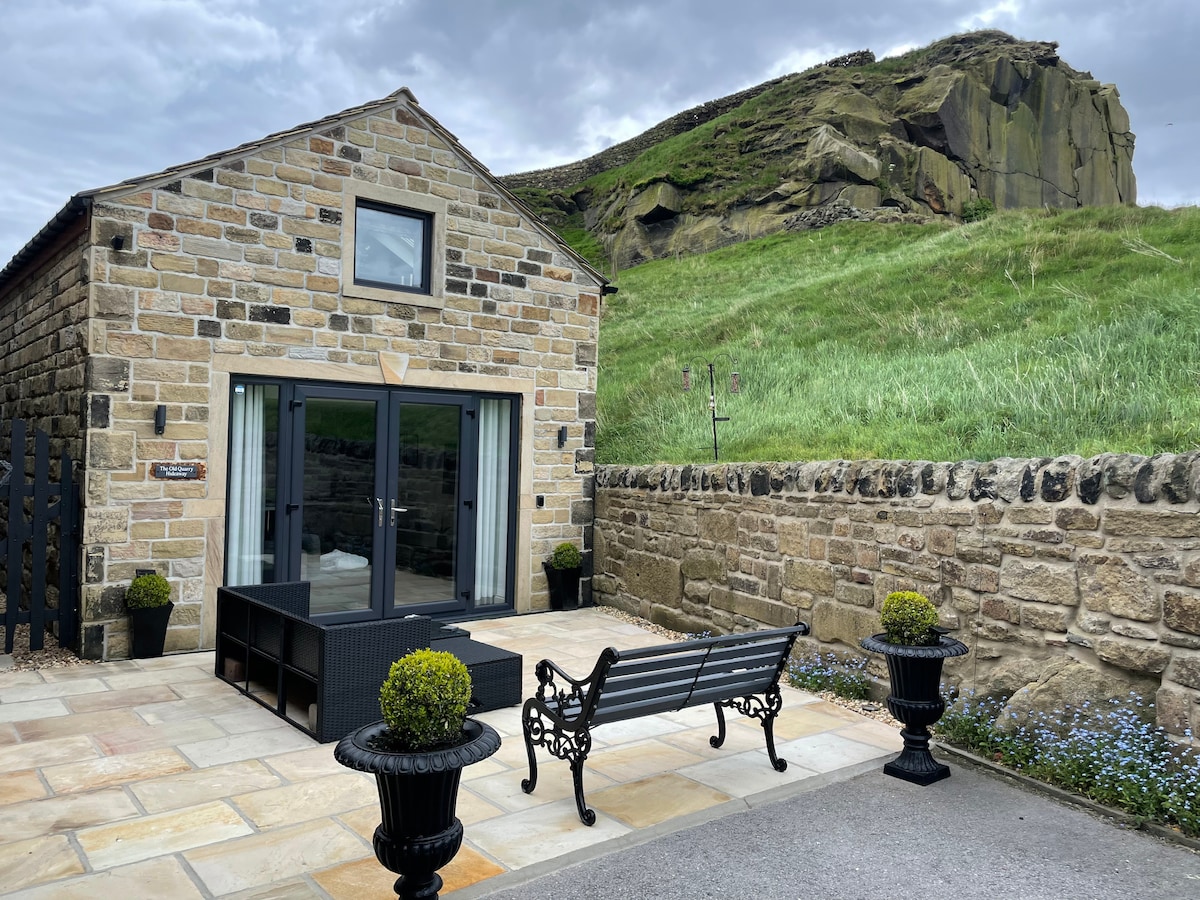 An exterior view showcases a stone cottage with large glass doors opening to a patio area. Two potted plants frame a decorative bench, and a backdrop of a grassy hillside creates a serene setting. Walled garden features natural stone and vibrant greenery.