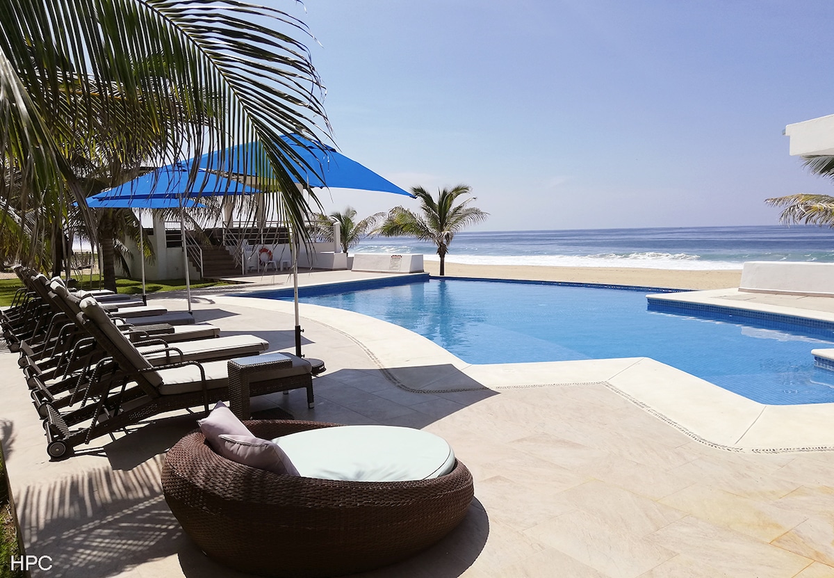 A serene pool area is set against a backdrop of ocean views. Lounge chairs are arranged around a large, inviting pool, complemented by shaded areas featuring blue canopies. Palm trees gently frame the scene, contributing to a calm and relaxing atmosphere.