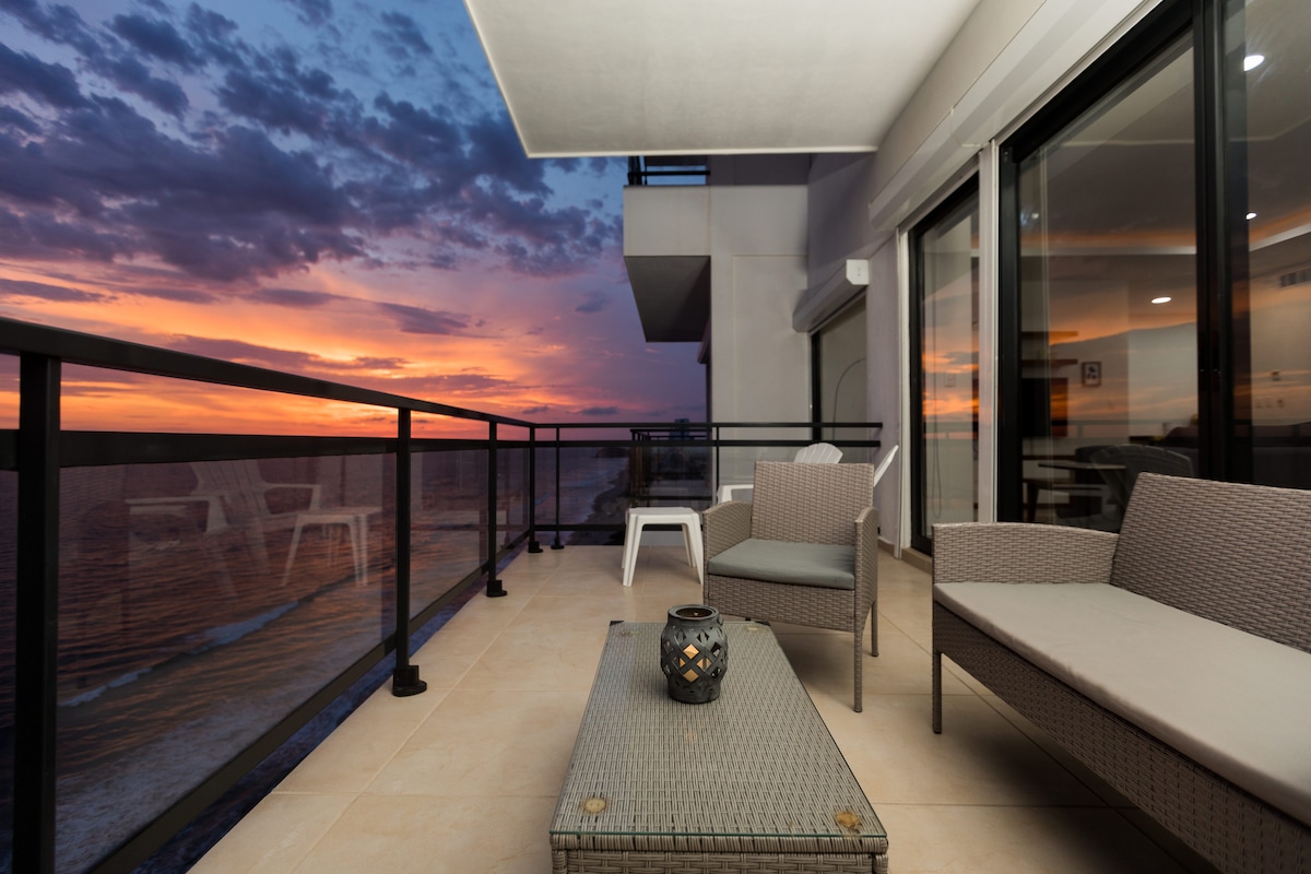 A spacious balcony offers a table and seating arrangements, featuring a modern design. A view of the sunset over the ocean is displayed, with vibrant colors illuminating the sky. A lantern sits atop the table, adding a cozy touch to the outdoor space.