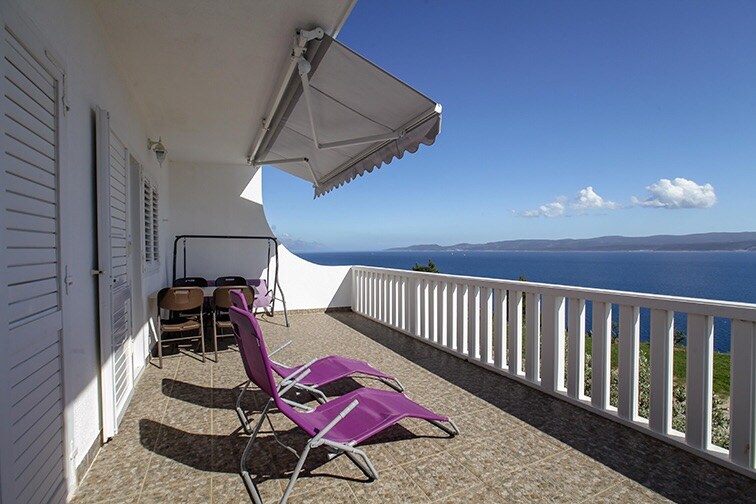 A private terrace is shown, featuring two purple lounge chairs positioned to overlook the sea. A white railing borders the space, while an awning provides shade. The expansive view includes clear blue water and distant mountains under a sunny sky.