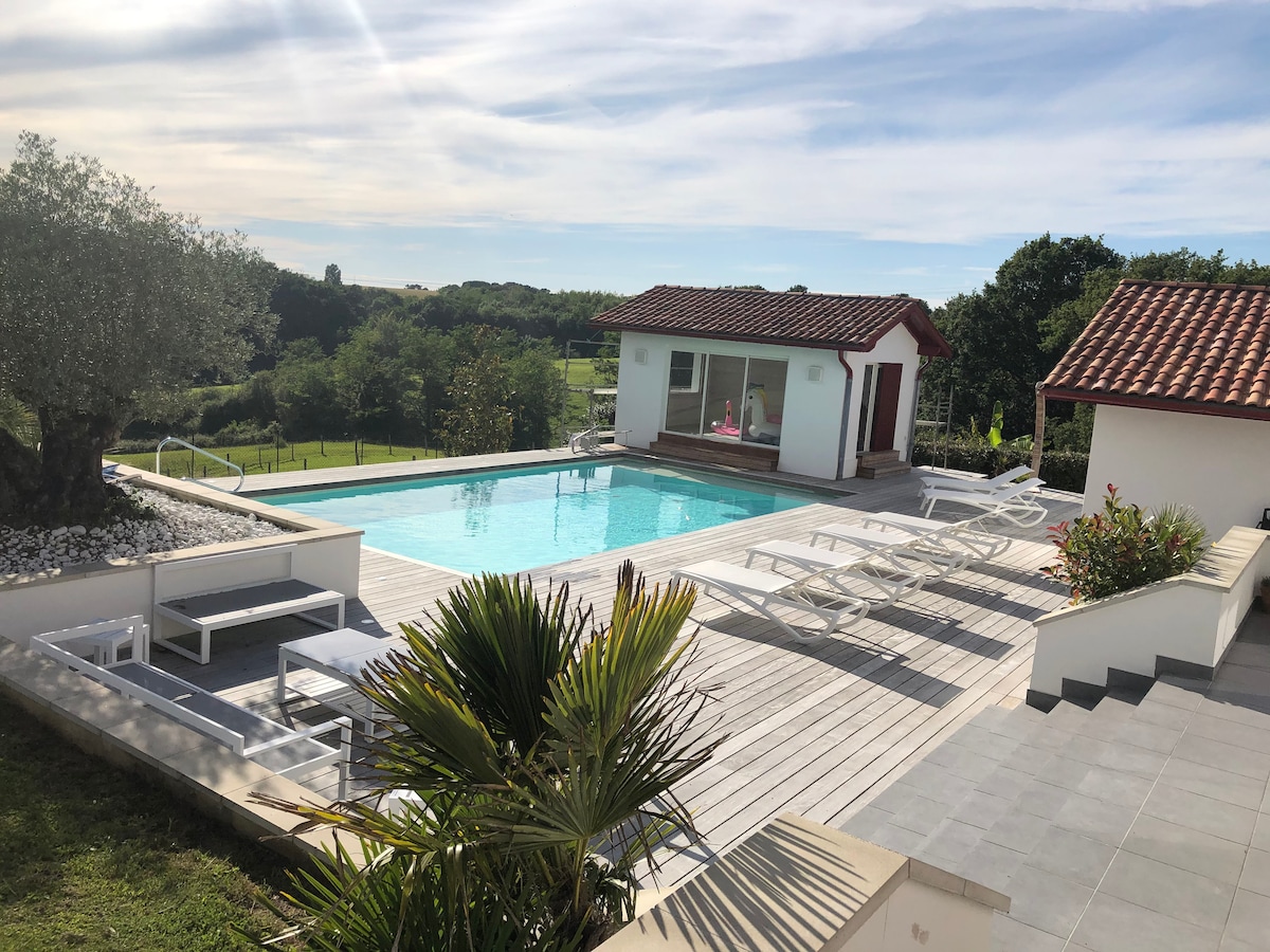 A spacious pool area is surrounded by greenery, featuring a heated swimming pool with a wooden deck. Several lounge chairs are positioned near the pool, while a charming building with large windows offers a view of the outdoor space.