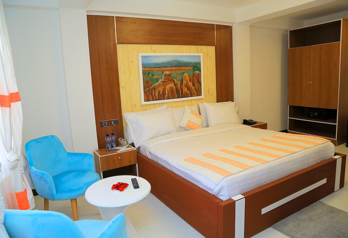 A spacious sleeping area is furnished with a large bed featuring a light-colored comforter. Vibrant orange accents are present in both the bedding and decorative elements. Two blue chairs are positioned next to a small wooden table, with a television unit visible in the background.