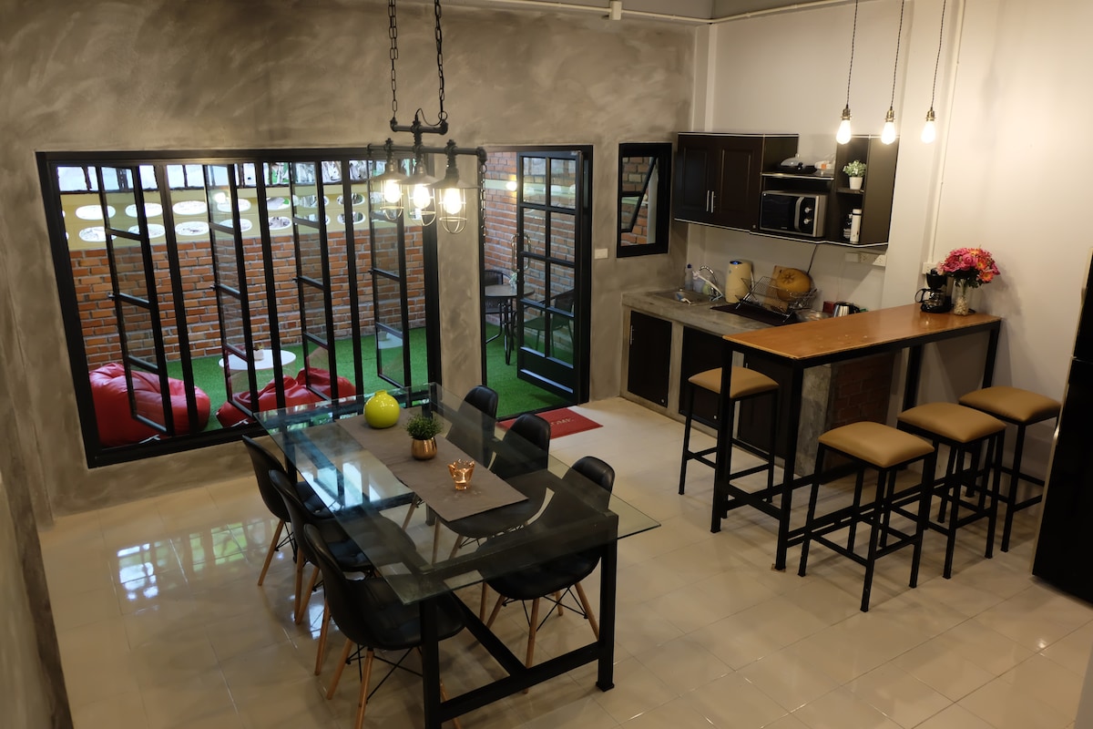 The dining area is highlighted by a large glass table surrounded by black chairs. An industrial-style bar with high stools complements the space. Large windows allow natural light to fill the area, while greenery can be seen outside, creating a bright and open atmosphere.