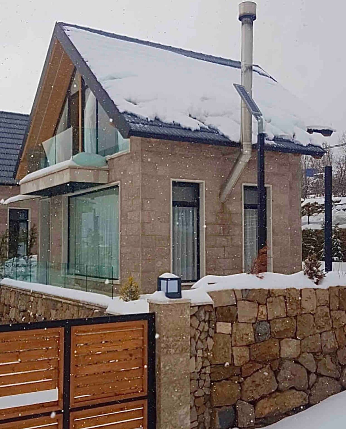 A modern chalet features large glass windows, allowing for natural light to fill the interior. Snow covers the rooftop, while a stone wall and wooden gate provide privacy. A chimney is visible, indicating a cozy atmosphere during winter months.