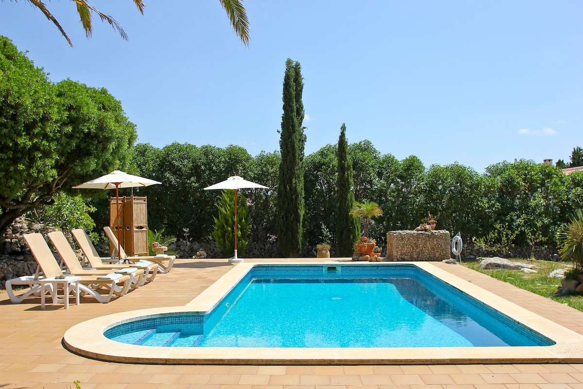 A private outdoor space features a swimming pool surrounded by stone tiles. Sun loungers with umbrellas are positioned nearby, creating a relaxing environment. Lush greenery provides privacy, and garden elements can be seen in the background, enhancing the serene setting.