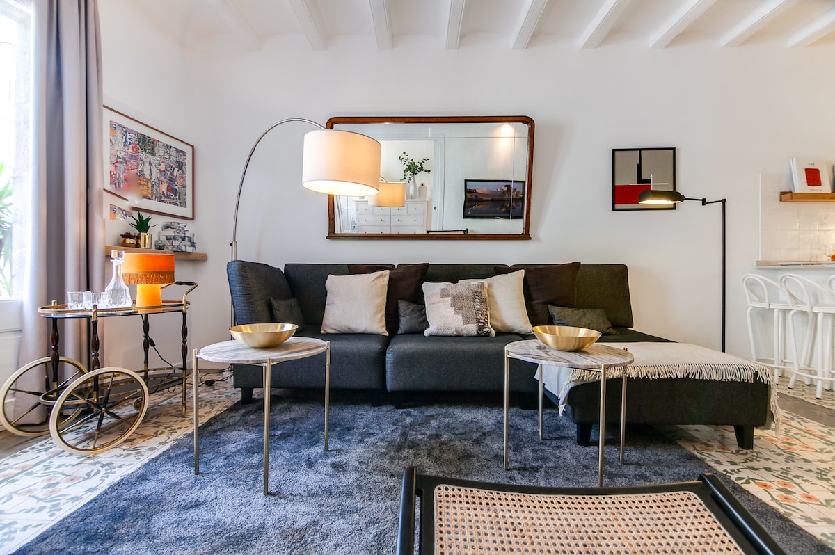 A sofa with neutral-colored cushions is arranged in a living area, complemented by two small side tables. A curved floor lamp and a wall-mounted mirror add functionality and style. Natural light enters through a nearby window, illuminating the patterned tile floor.