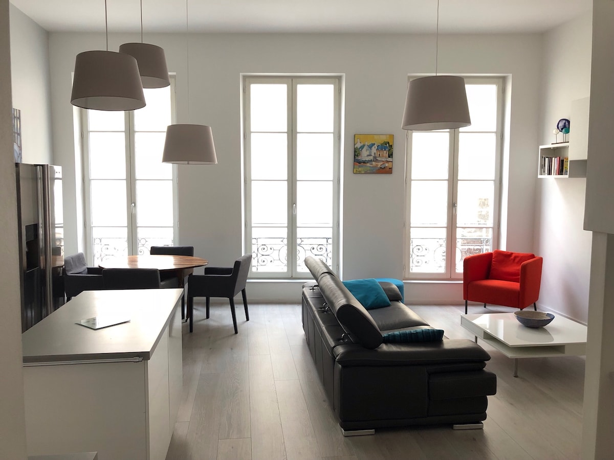 A spacious living area features large windows that invite natural light, complemented by a mix of modern furniture. A leather sofa is positioned with colorful cushions, alongside a sleek coffee table. A wooden dining table is visible, seating four, with adjacent chairs and decorative elements on shelves.