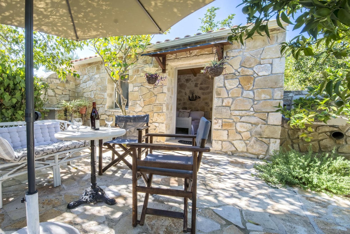 A covered terrace is set with outdoor dining furniture, surrounded by lush greenery and stone walls. Natural light filters through the overhead leaves, creating a serene space. The entrance to the studio house is visible in the background, enhancing the private ambiance of the setting.