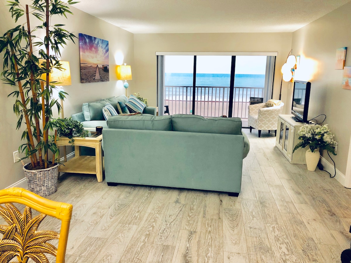 A spacious living area is highlighted by large windows offering ocean views. A comfortable teal sofa and armchair are arranged around a coffee table. Indoor plants and decorative items enhance the decor. Soft lighting from mounted wall fixtures adds ambiance to the space.