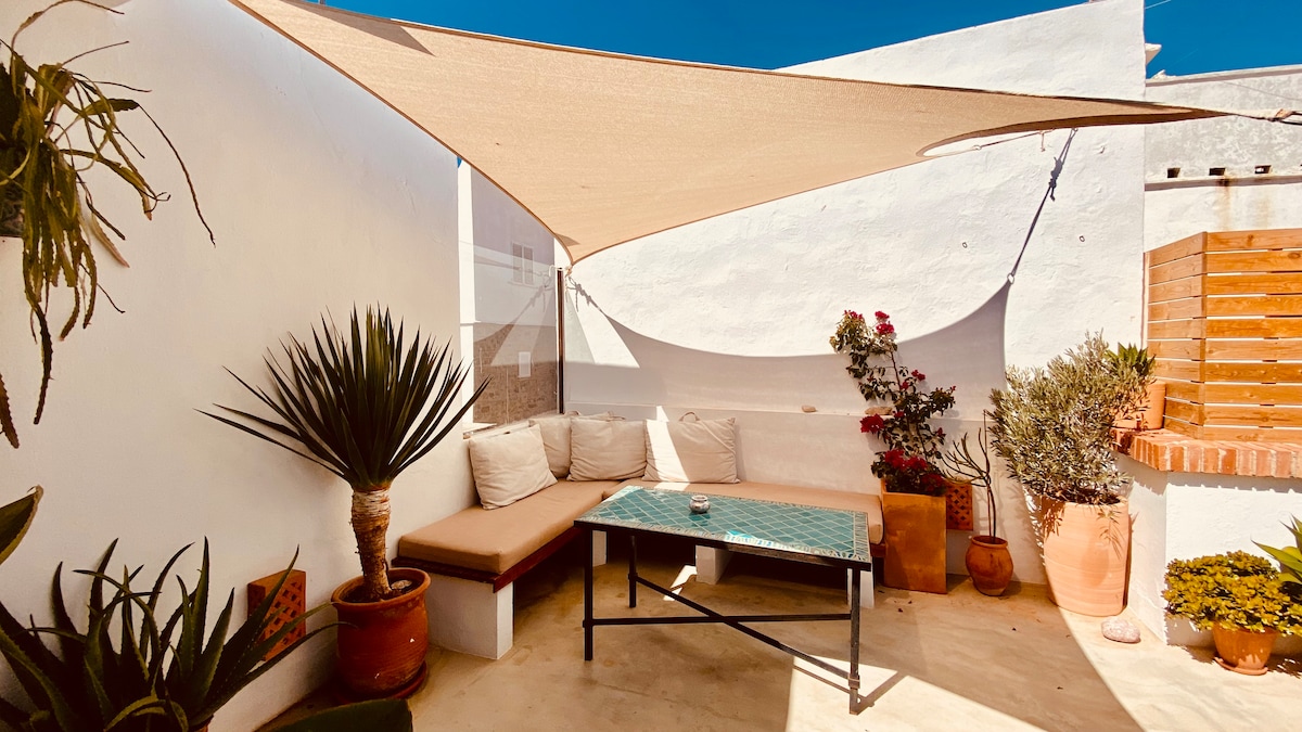 A private terrace is presented, featuring a sunshade overhead. A comfortable seating area with light-colored cushions surrounds a table. Potted plants, including a tall palm and flowering shrubs, add greenery to the space, while the walls are clad in white, creating a bright and airy atmosphere.