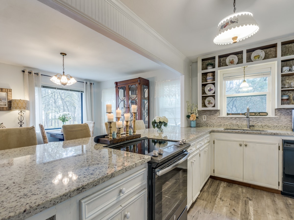 A spacious kitchen with light-colored cabinetry and granite countertops is depicted. An elegant chandelier offers warm illumination. A dining area with large windows and tasteful decor is visible through an open space, enhancing the flow between areas while natural light brightens the room.