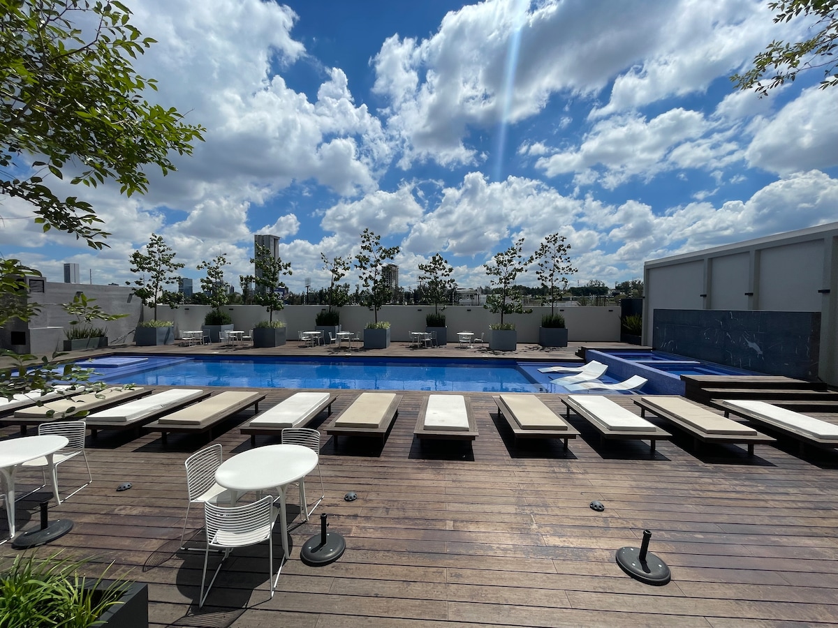 A serene pool area is surrounded by lounge chairs and small tables, offering a relaxing outdoor space. Lush greenery and modern planters frame the setting, while the bright blue sky is dotted with fluffy clouds overhead.