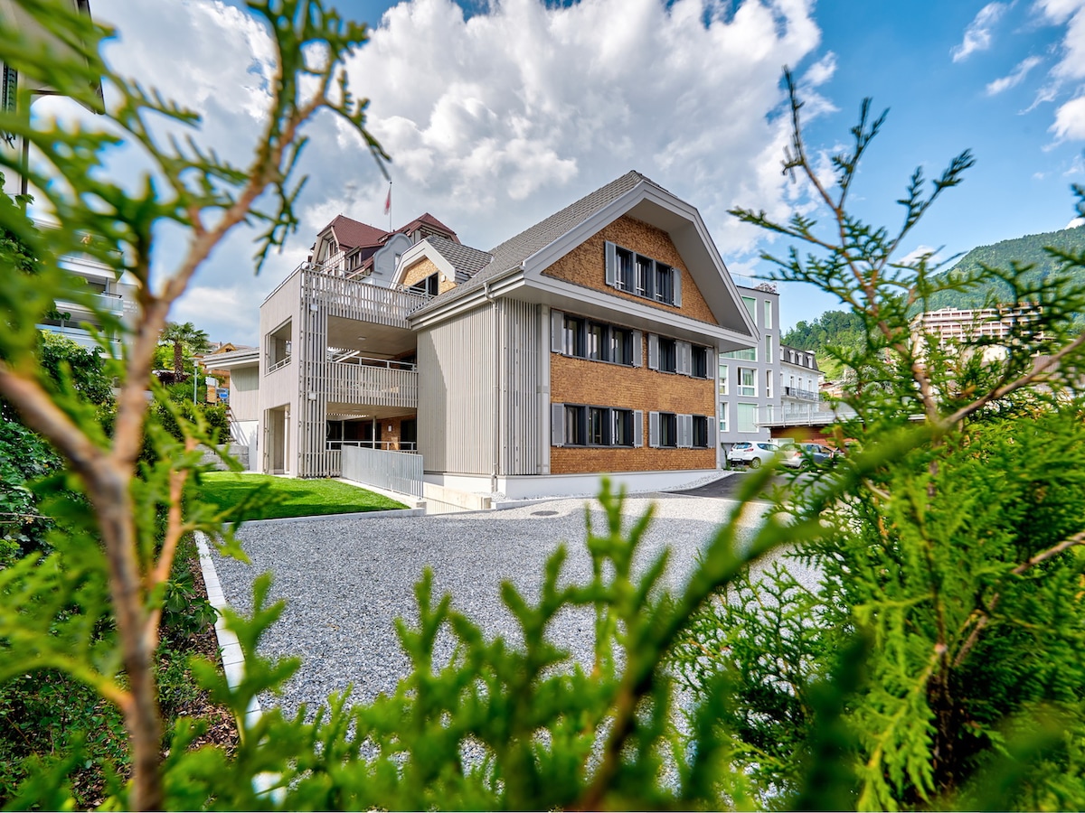 The guesthouse is showcased from a greenery-framed perspective, presenting its modern facade with a blend of traditional and contemporary elements. The structure features multiple windows, allowing natural light to fill the interior, with a gravel pathway leading to the entrance.