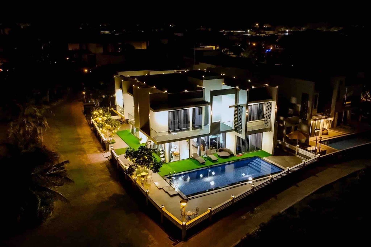 The villa is showcased at night, illuminated by soft outdoor lighting. A large pool is surrounded by greenery and loungers, creating a serene ambiance. The spacious structure features balconies and large windows, providing views of the surrounding landscape.