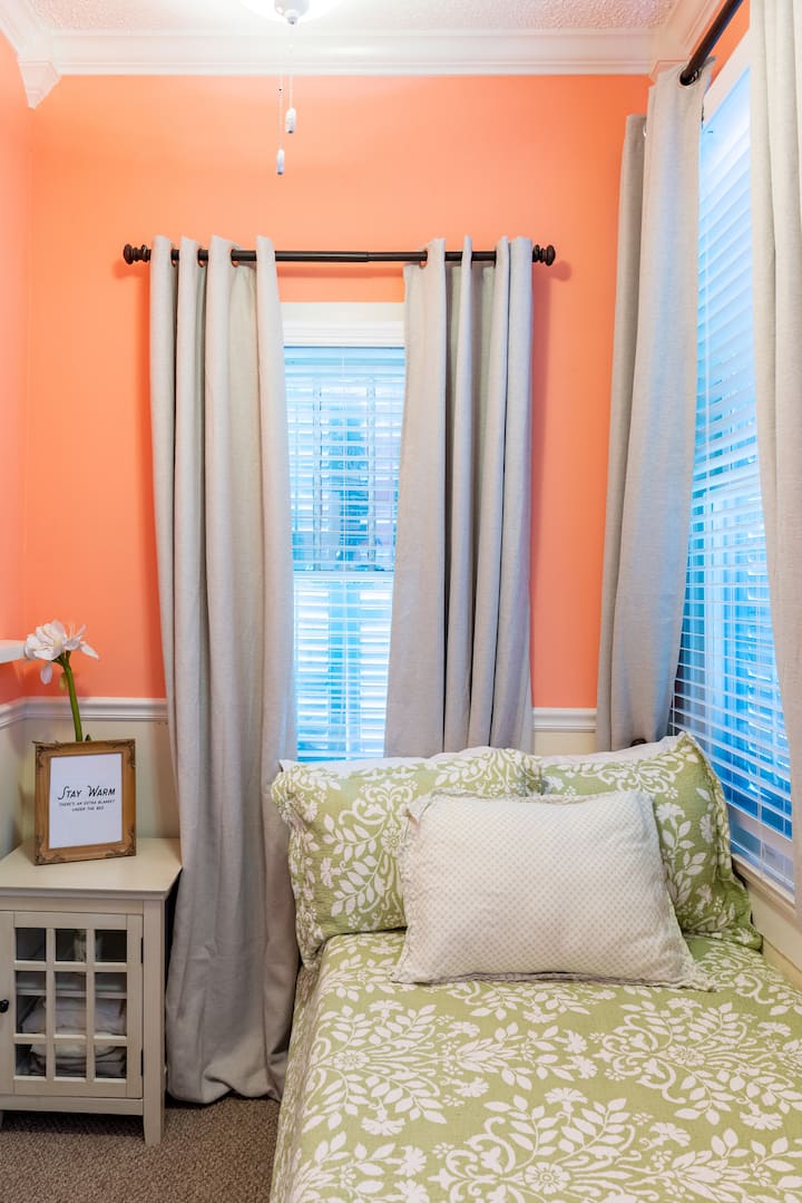 - חדר שינה נעים עם קירות קורל עליזים ואור טבעי משני חלונות
- מצעים פרחוניים ירוקים ולבנים עם כרית נוי גדולה מוסיפים נוחות
- על השידה ליד המיטה יש יצירות אמנות ממוסגרות ופרח אחד שמעניק מגע חם