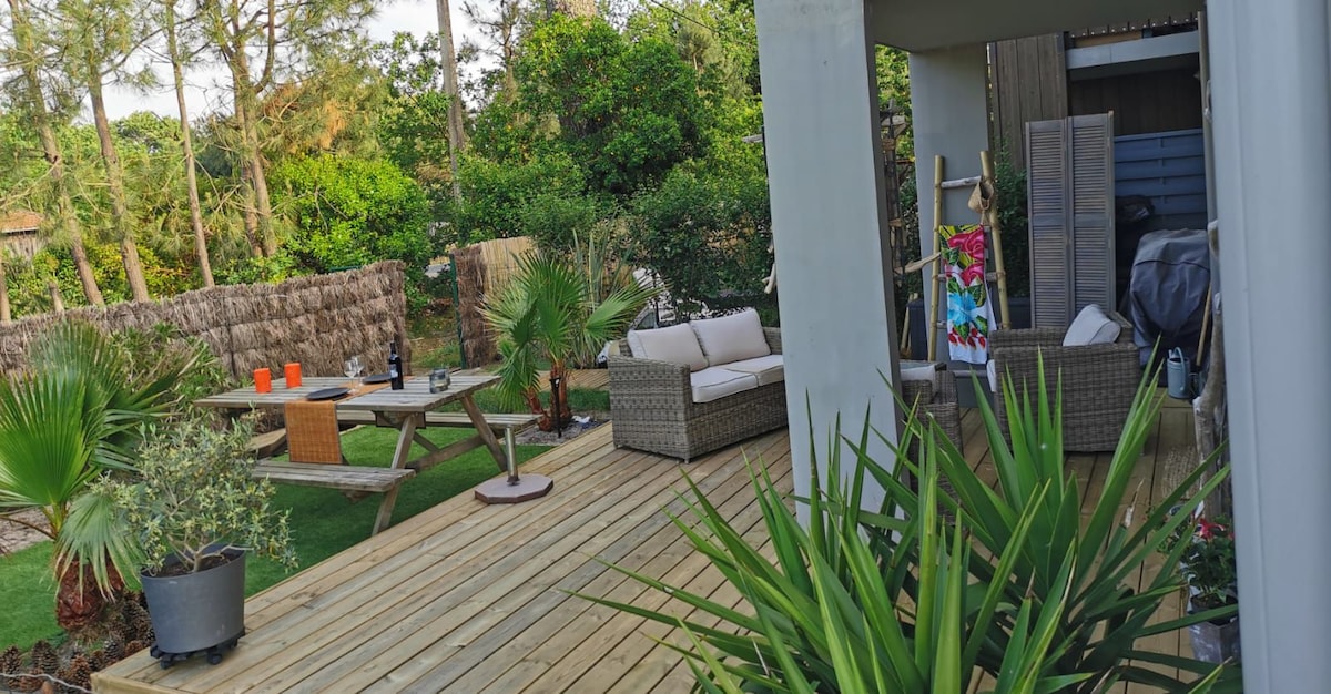 A spacious wooden terrace features a comfortable seating area with a sofa and coffee table. A dining setup with orange chairs is visible in a lush garden surrounded by palm trees and other greenery. The atmosphere reflects a calm outdoor space designed for relaxation.