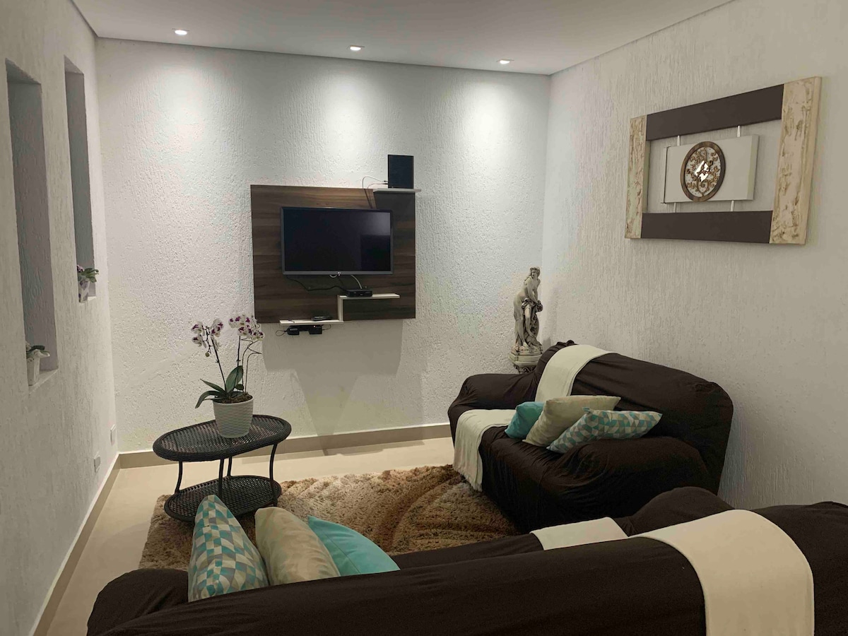 A comfortable living area is arranged with two brown sofas adorned with light-colored cushions. A flat-screen TV is mounted on the wall above a dark wooden media unit. A circular table is positioned between the sofas, topped with decorative elements and surrounded by a soft area rug.