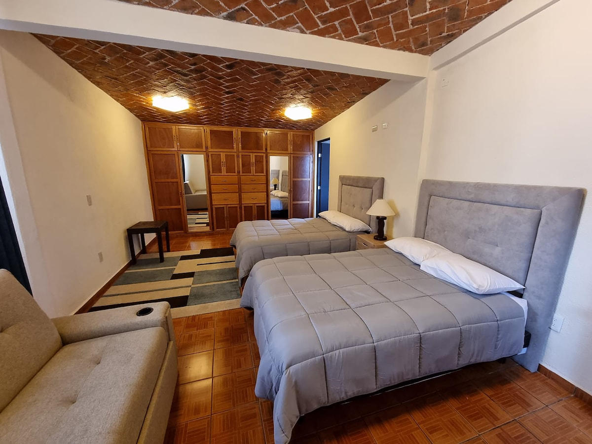 A spacious room is furnished with two beds topped with gray bedding. A light-colored sofa sits in the corner beside a patterned rug. A wooden wardrobe and dressing area reflect natural light coming from a nearby window.