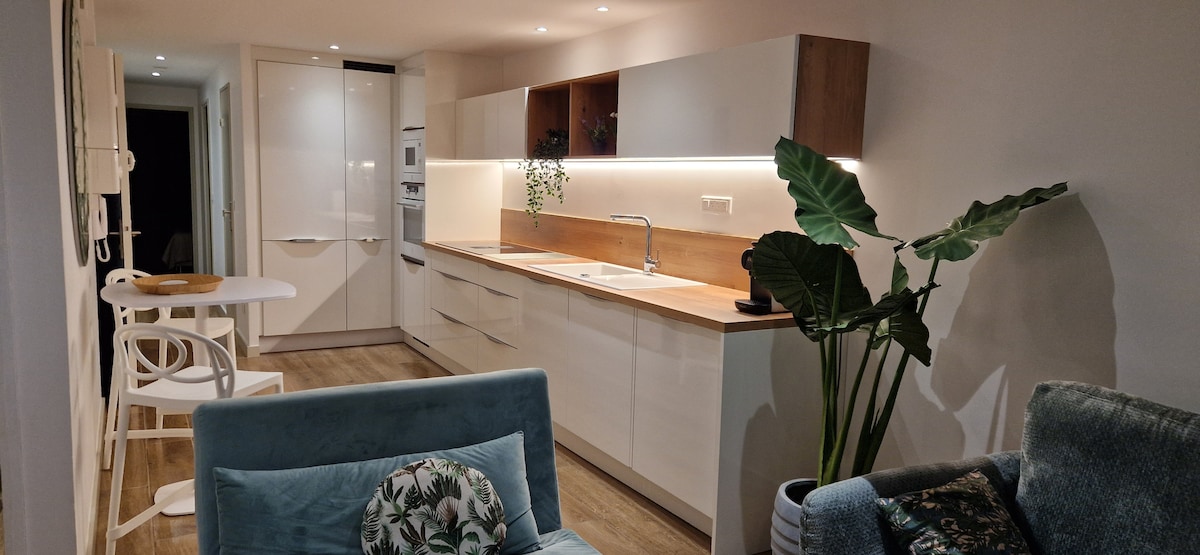 A modern kitchen area features sleek white cabinetry with wooden accents. A countertop offers space for meal preparation, while a dining table with two chairs is positioned nearby. Indoor plants add a touch of greenery, contributing to the overall fresh ambiance.