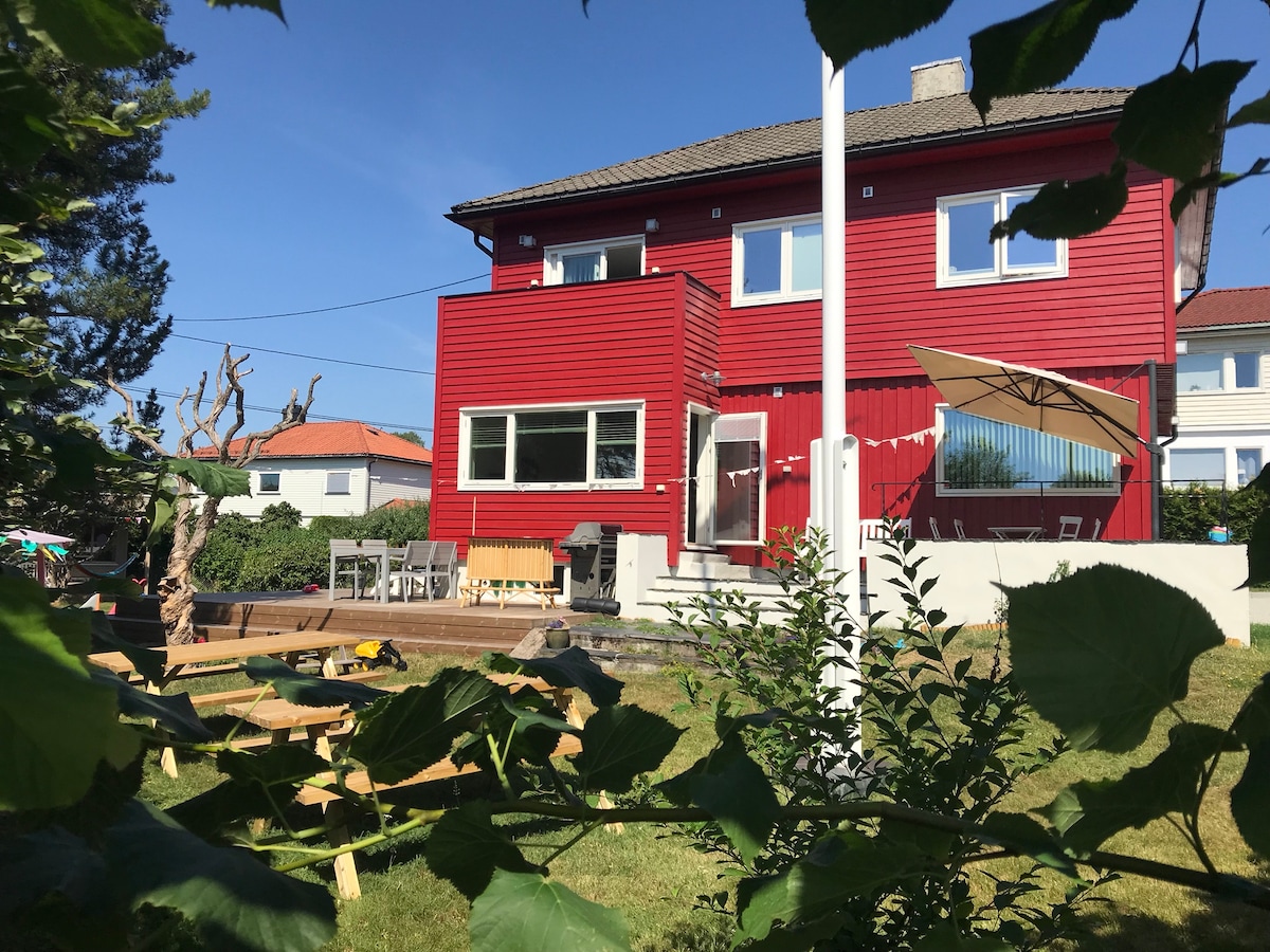 A classic red home is viewed from the garden, showcasing large windows and a welcoming entrance. An outdoor area with seating is visible, surrounded by lush greenery. The clear sky above enhances the appealing environment of the spacious yard.